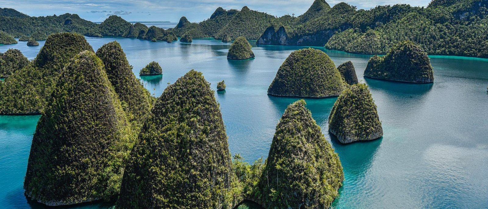 Steep green karst islets rising from clear turquoise water at Wayag in Raja Ampat, with more forested islands and a bright sky beyond.