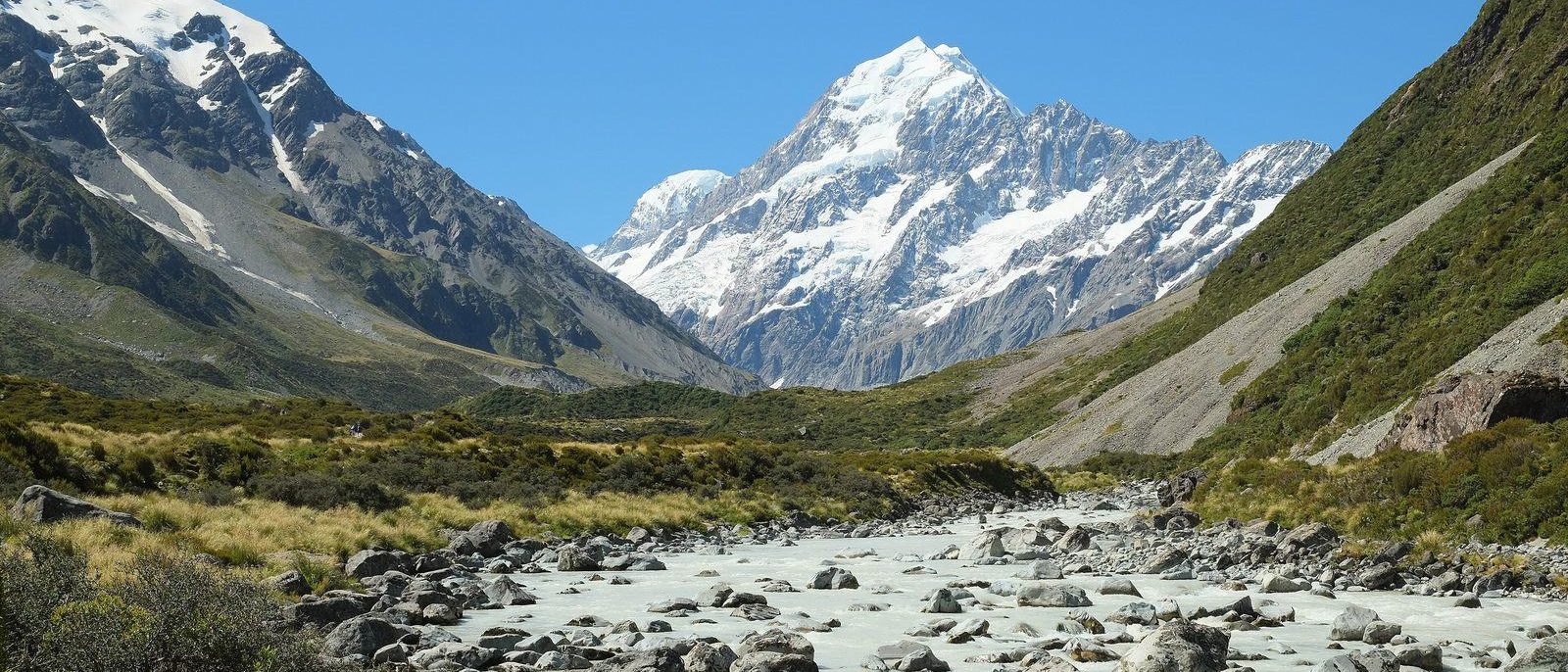 Aoraki / Mount Cook, its snow-covered summit rising at the head of the Hooker Valley above a braided, milky glacial river and a boulder-strewn riverbed.
