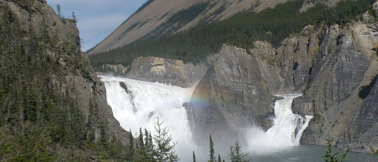 Virginia Falls on the South Nahanni River, white water pouring around a central rock pillar into a canyon, with spruce in the foreground and a forested mountainside behind.