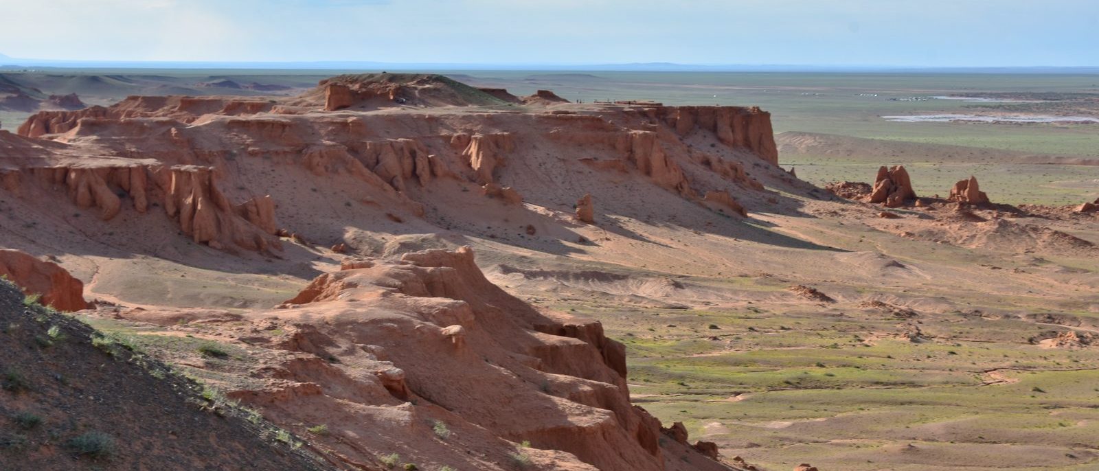The Flaming Cliffs at Bayanzag: red-orange sandstone escarpments carved into buttresses and gullies, above the flat green-brown floor of the Gobi stretching to the horizon.