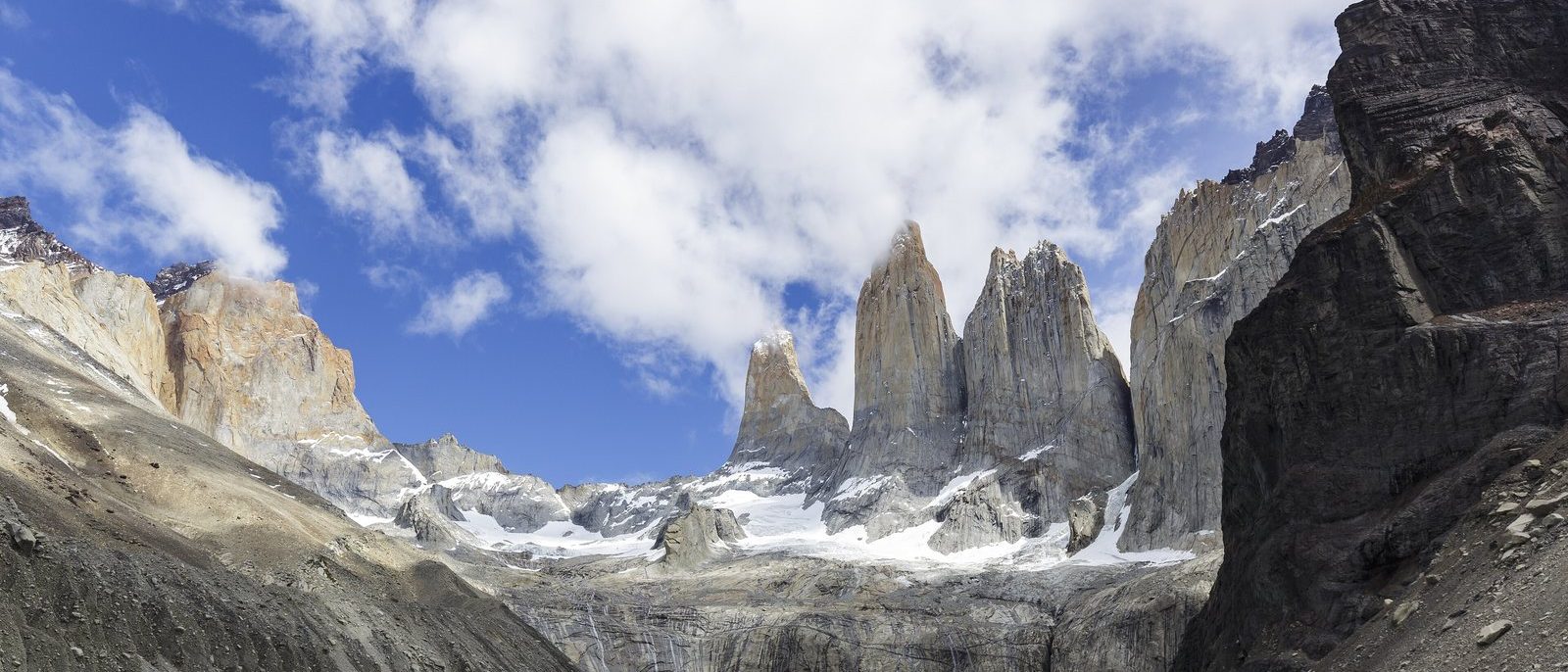 The granite towers of Torres del Paine rising above a turquoise glacial lake under a blue sky of white cloud, a dark rock wall at the right and pale boulders in the foreground.