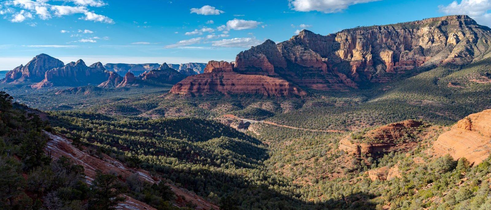 Red sandstone buttes and mesas above a wide valley of juniper and pine near Sedona, with slickrock in the foreground and white clouds in a blue sky.