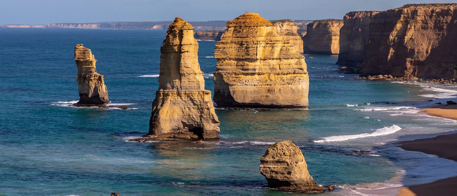 Limestone sea stacks of the Twelve Apostles standing in blue water off the Victorian coast, lit gold by low sun, with cliffs running away to the right.