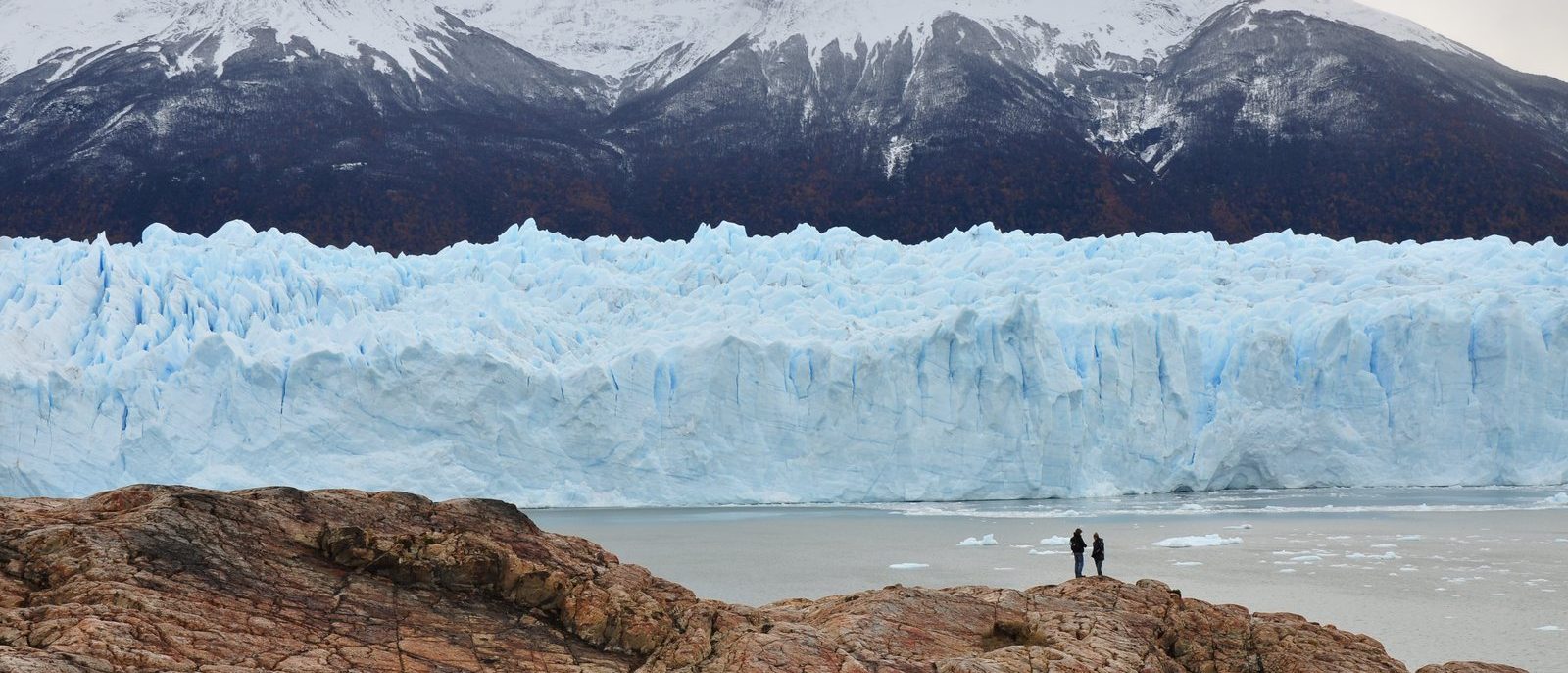 The pale blue ice front of the Perito Moreno Glacier stretching across the frame beneath snow-covered mountains, two people standing tiny on a rock outcrop in the foreground.
