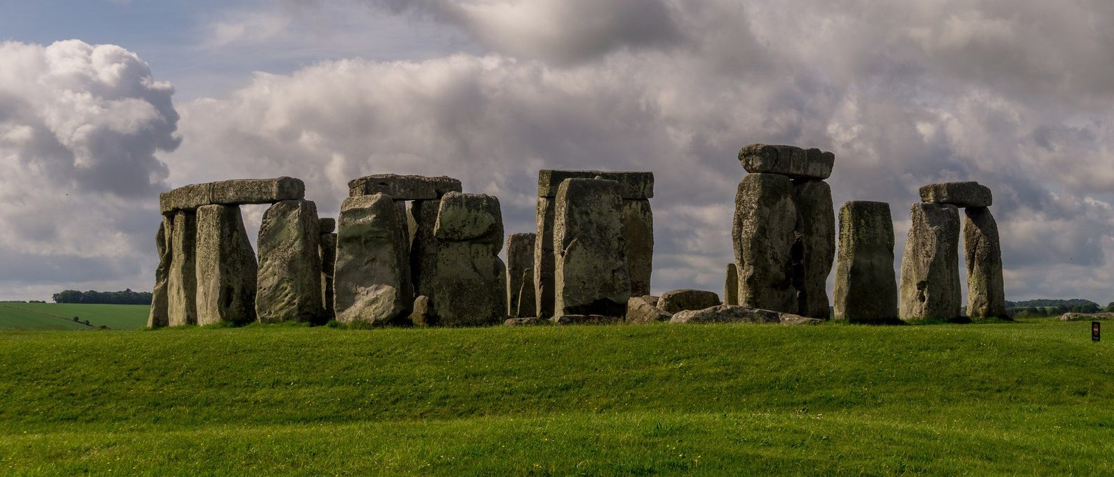 The standing stones of Stonehenge on green grass beneath a broad cloudy sky, lintels still capping several of the uprights.