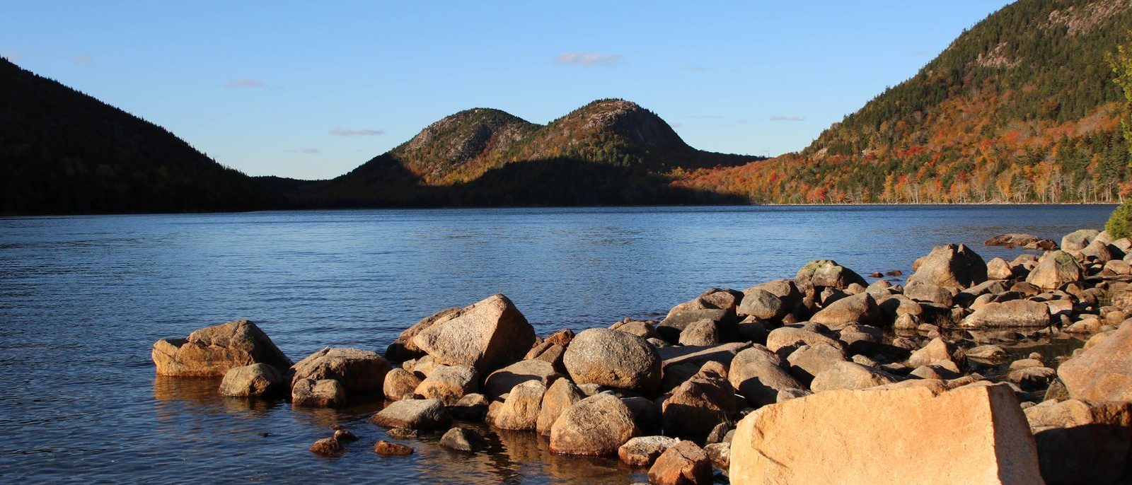 Jordan Pond in Acadia National Park in late sun, granite boulders along the near shore and the two rounded hills called the Bubbles beyond, their slopes turning autumn orange.