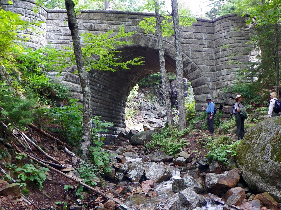 A rough-faced granite arch bridge over a wooded path