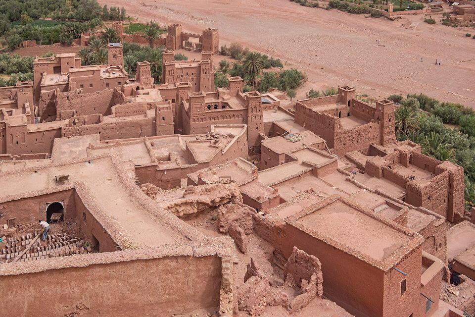 Flat red-brown earthen rooftops and towers seen from a height, palms and green fields beyond, rows of mud bricks laid out at the lower left
