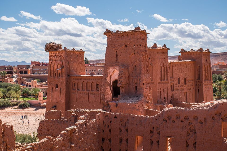 Red-brown earthen towers with patterned tops and a gateway arch in hard sun under a blue sky with clouds, a stork's nest on the left-hand tower, the newer village beyond and a few tiny walkers in the riverbed