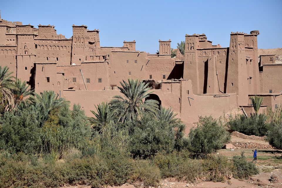 Earthen tower houses and walls of the ksar rising above palms and scrub on the riverbank under a clear blue sky, a small figure in a blue robe walking at the lower right
