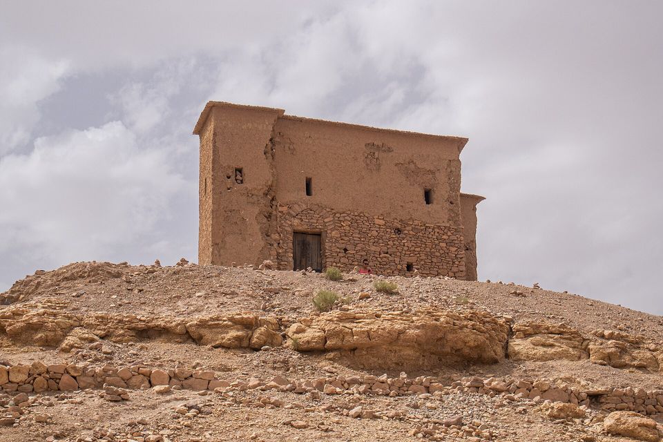 A flat-roofed building of stone below and earth above, with a wooden door, standing on a rocky mound under a cloudy sky