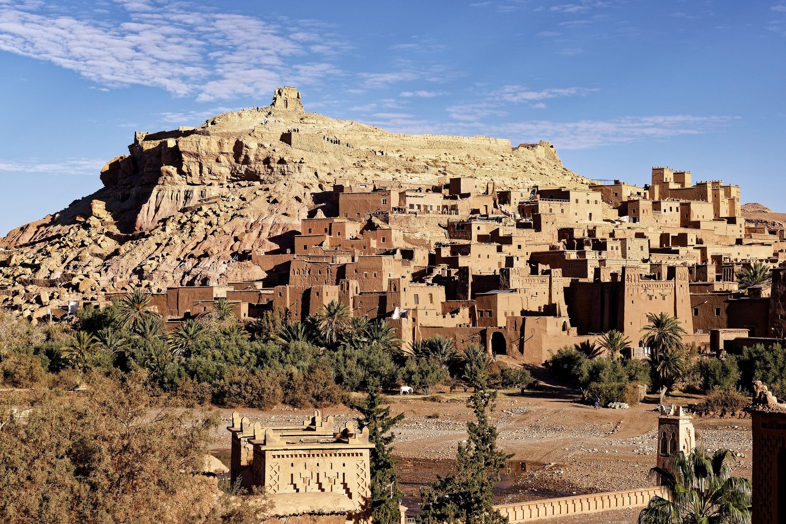 A hillside of flat-roofed earthen houses and towers rising to a ruined structure on a rocky summit, palms and a dry riverbed in front, under a blue sky with streaks of cloud