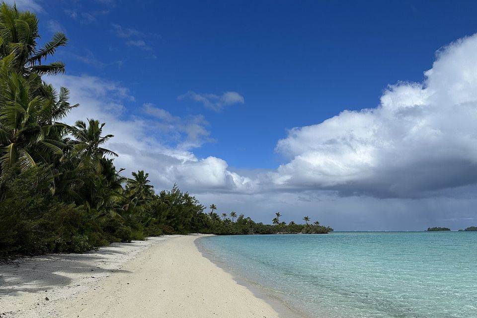 A curve of white sand under leaning palms on Motu Akaiami, with the lagoon's shallows stretching to small islands on the horizon