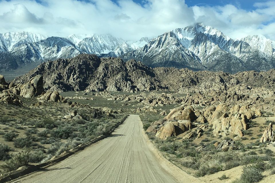 A dusty dirt road crossing a plain of granite boulders with the snowy Sierra Nevada under clouds behind