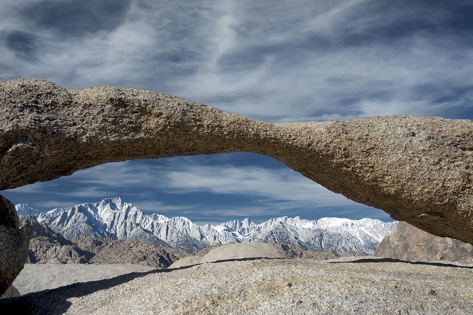 A long low granite arch framing a snowy mountain crest under streaky cloud