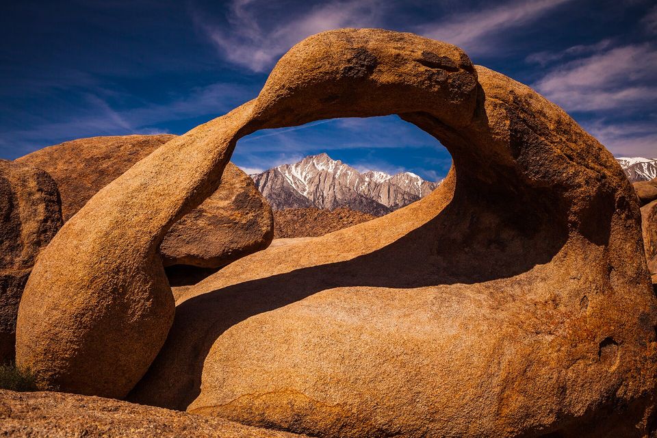 A natural granite arch in orange boulders with the snowy Sierra Nevada seen through the opening