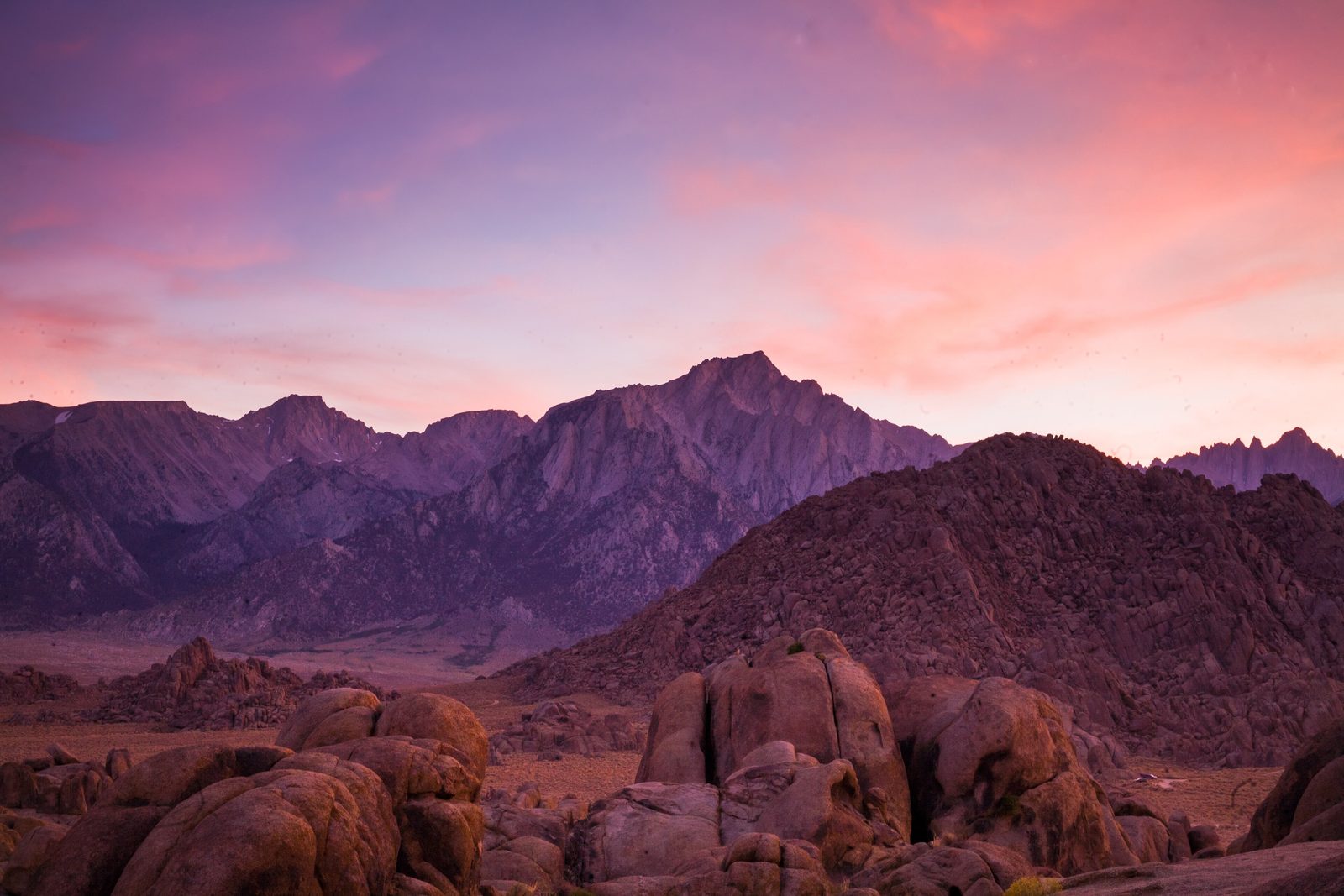 Rounded orange granite boulders in the foreground, a dark boulder hill behind and the Sierra Nevada wall under a pink and violet dusk sky