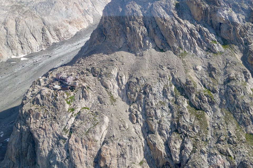 An aerial view of a small mountain hut perched on a rocky spur high above a grey, debris-covered glacier