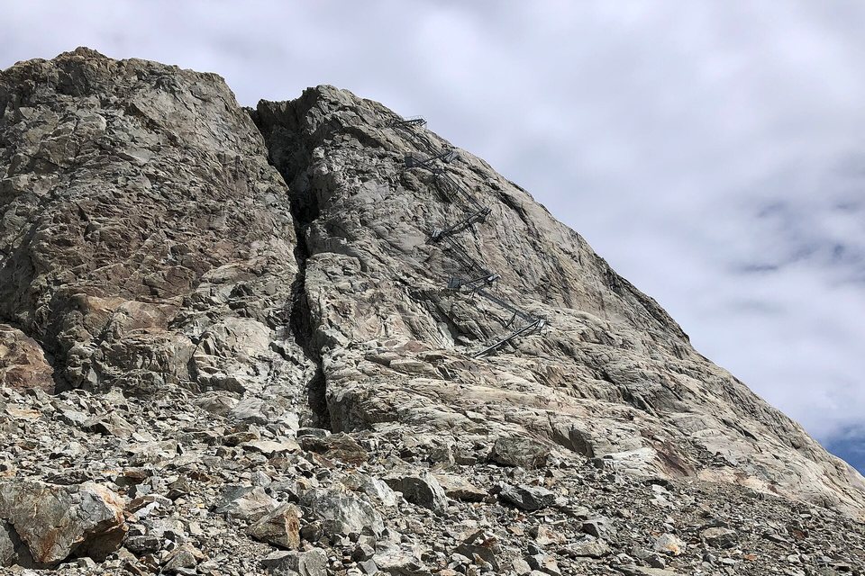 A steep pale rock face above a scree slope, with a zigzag line of metal staircases bolted up it