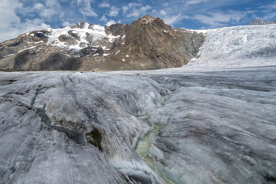 A pale green meltwater stream cut into grey glacier ice, a rocky peak with snow patches behind