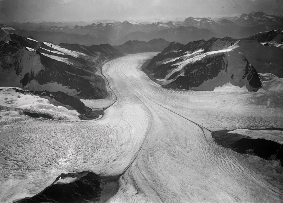 A black-and-white aerial photograph of a broad glacier with dark stripes of moraine sweeping between snowy ridges