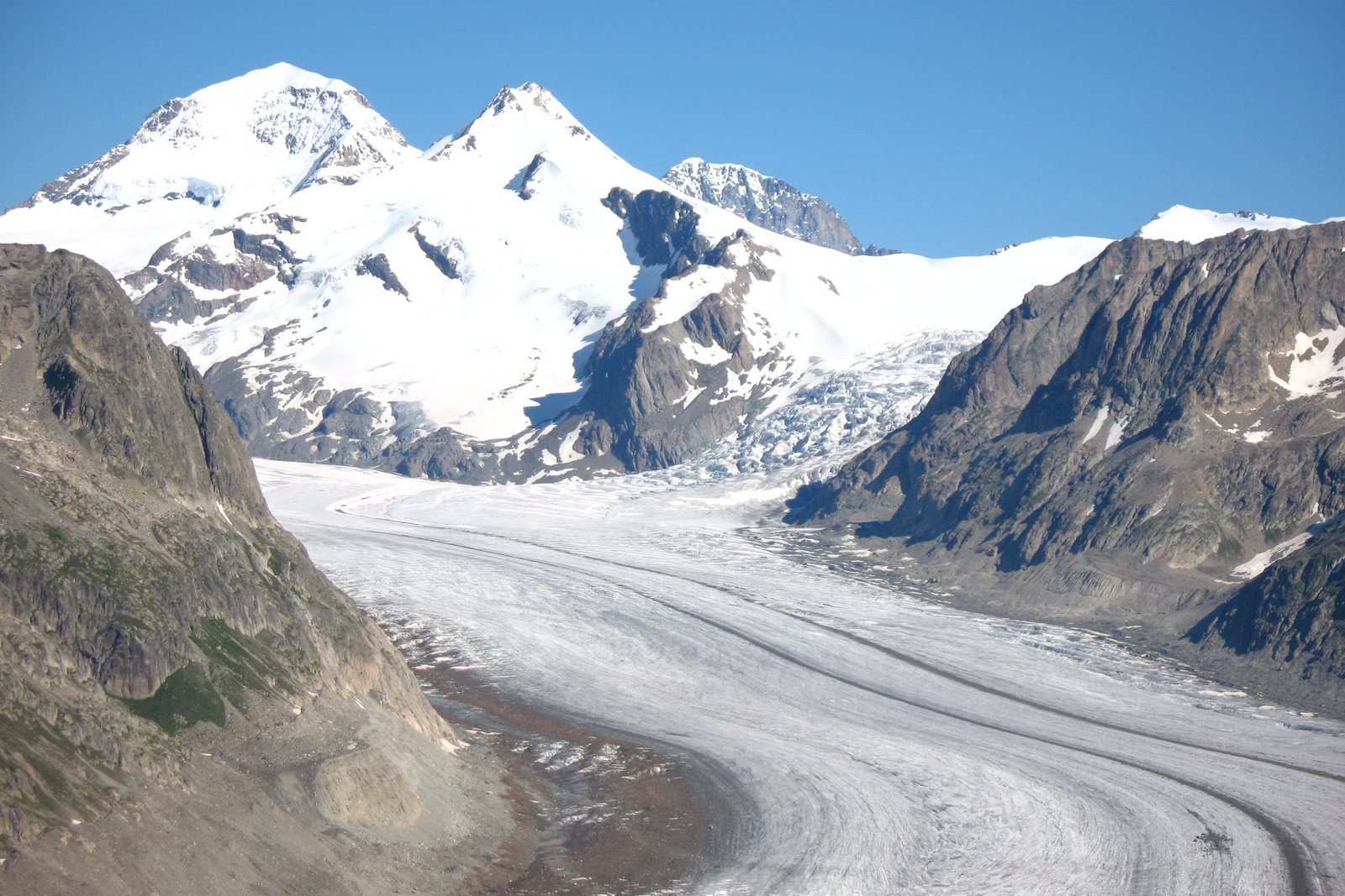 A broad grey-white glacier curving down a valley, long dark stripes of moraine running its length, snow peaks at its head