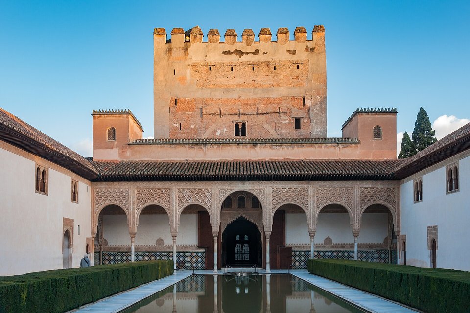 A long rectangular pool in a courtyard reflecting a tall sandstone tower above an arcade of carved arches, a hedge along each side, blue sky and cypresses behind