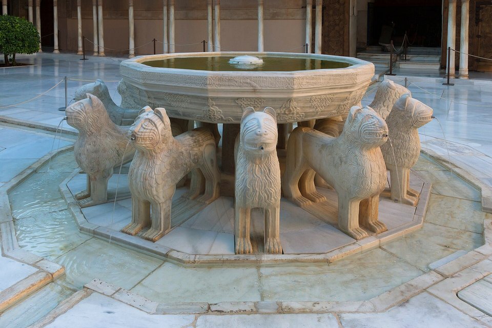 A wide stone basin on twelve lions standing in a paved court, water in the basin and thin jets running in channels from the lions’ mouths