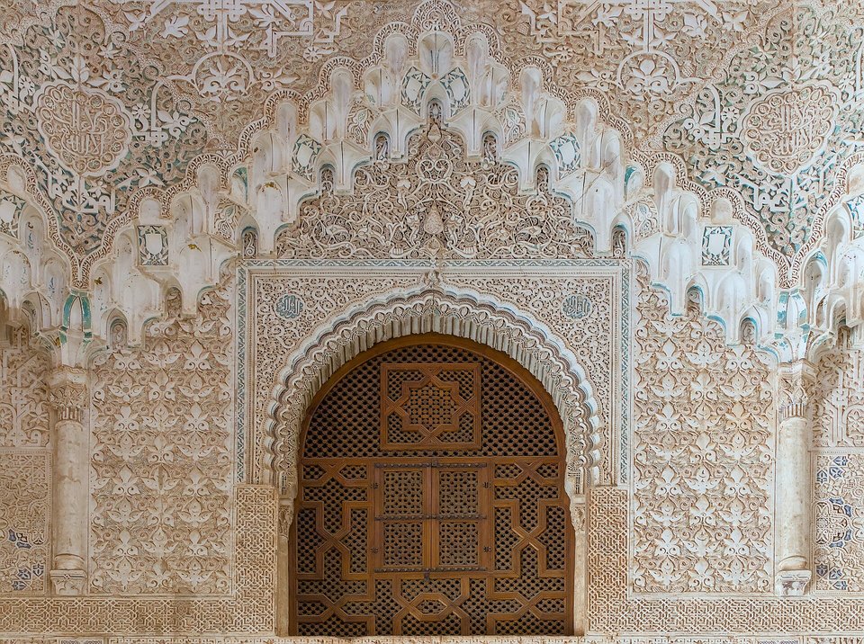 A tall arched window with a wooden lattice set in a wall of finely carved white plaster, honeycomb vaulting above it and coloured tile below