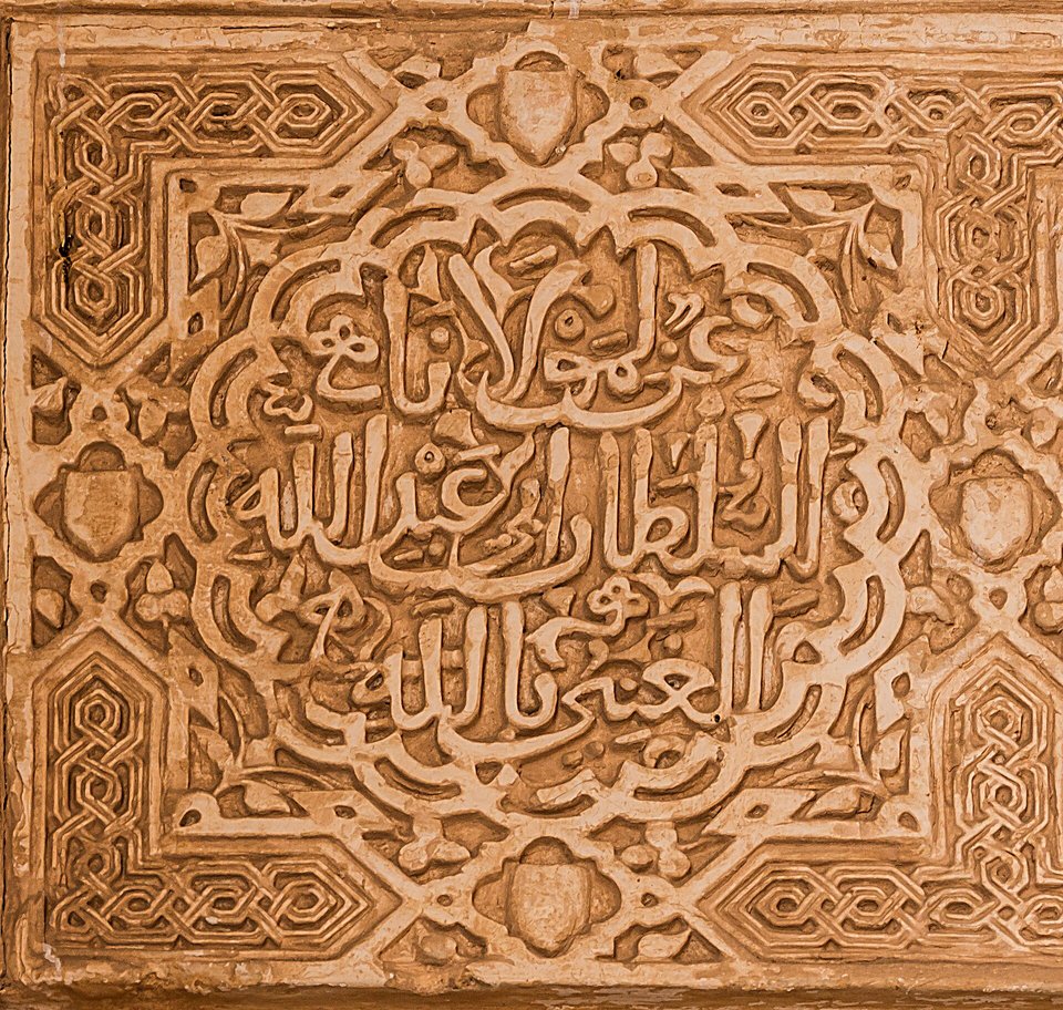 A square panel of carved plaster with Arabic script in raised letters framed by interlaced geometric borders, in warm sandy colour