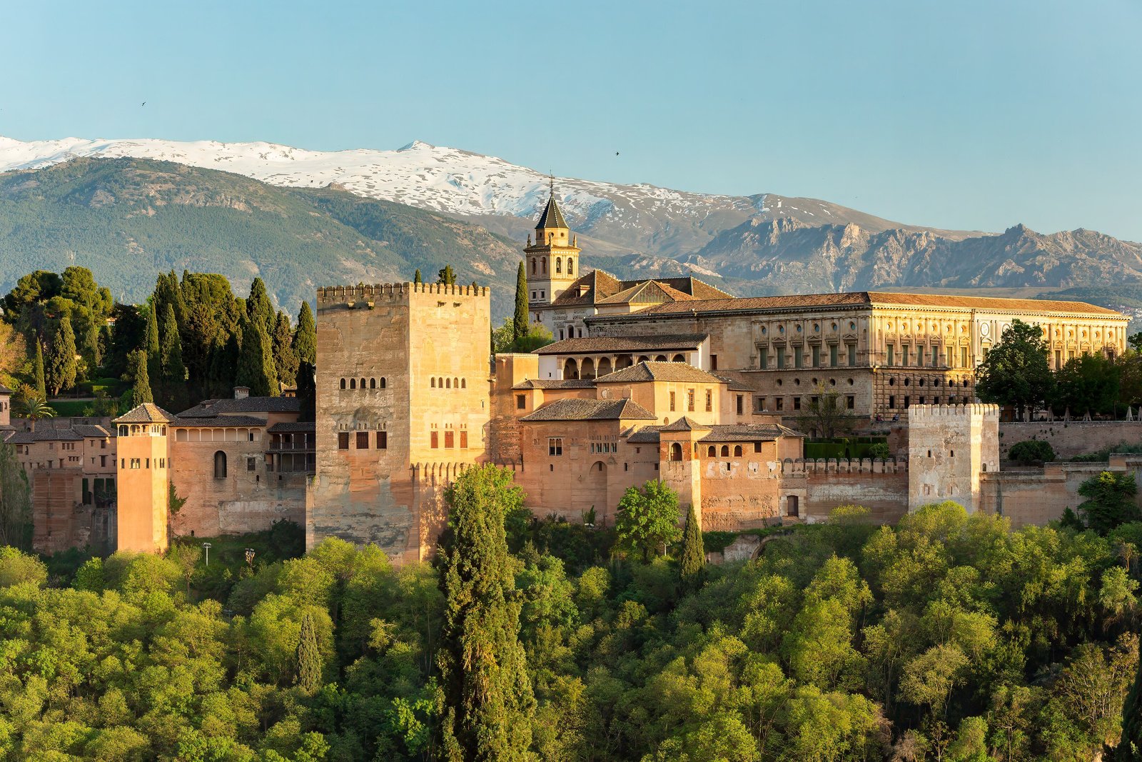 A hilltop complex of sandy-coloured walls, square towers and palaces in golden light above dense green trees, with a snow-capped mountain range behind