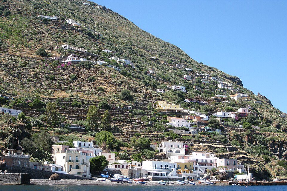 White and ochre houses step up a steep terraced slope above a pebble beach lined with small boats, under a clear blue sky