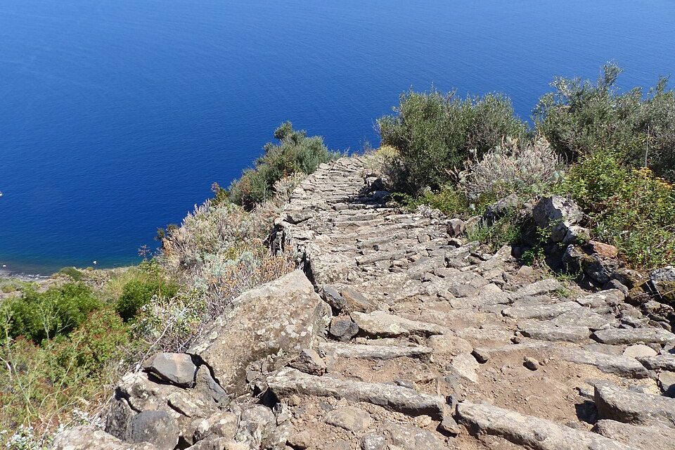 A rough stair of laid stone slabs runs down a scrubby hillside toward deep-blue sea far below