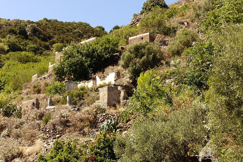 Roofless stone and whitewashed houses sit among olive scrub and prickly pear on a steep green slope