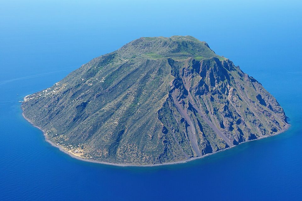 A steep volcanic island with gullied cliffs on one side and a few houses at the shore, in deep-blue sea