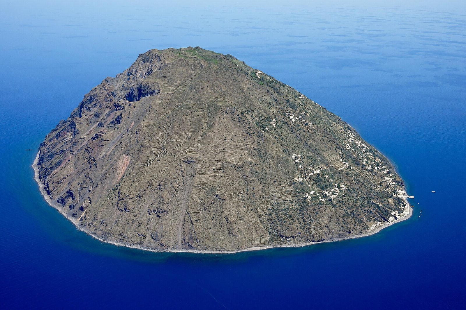 A steep, bare volcanic island seen from the air, its slopes ribbed with old terraces and white houses scattered down one side, in deep-blue sea