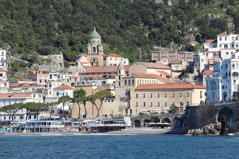 A town climbing a steep coast with a tiled cathedral dome, the coast road on arches beside the water