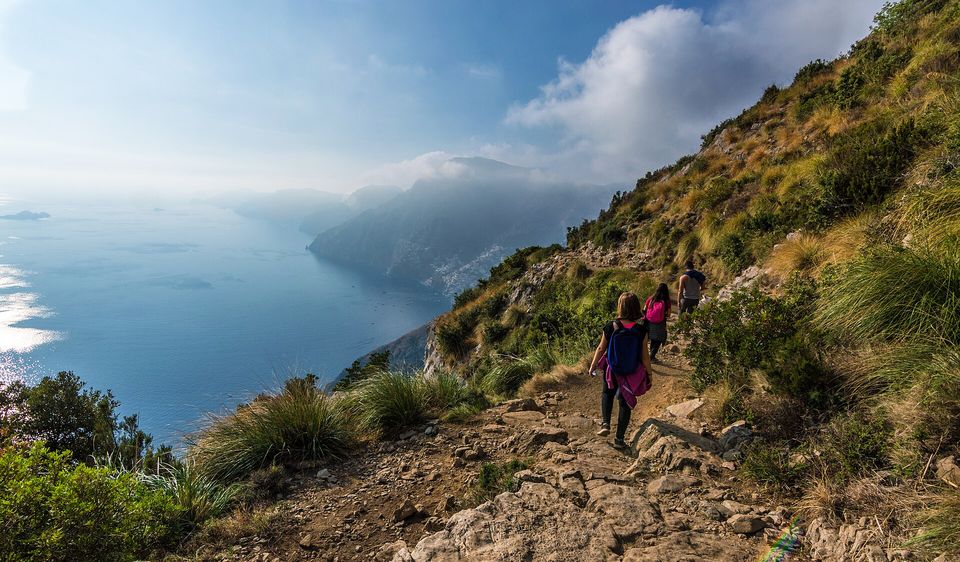 Walkers seen from behind on a stony path along a steep grassy hillside above a hazy blue coast