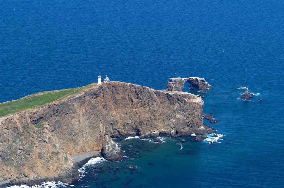 An aerial view of a steep brown headland topped by a white lighthouse, with a rock arch standing in deep blue water off its tip