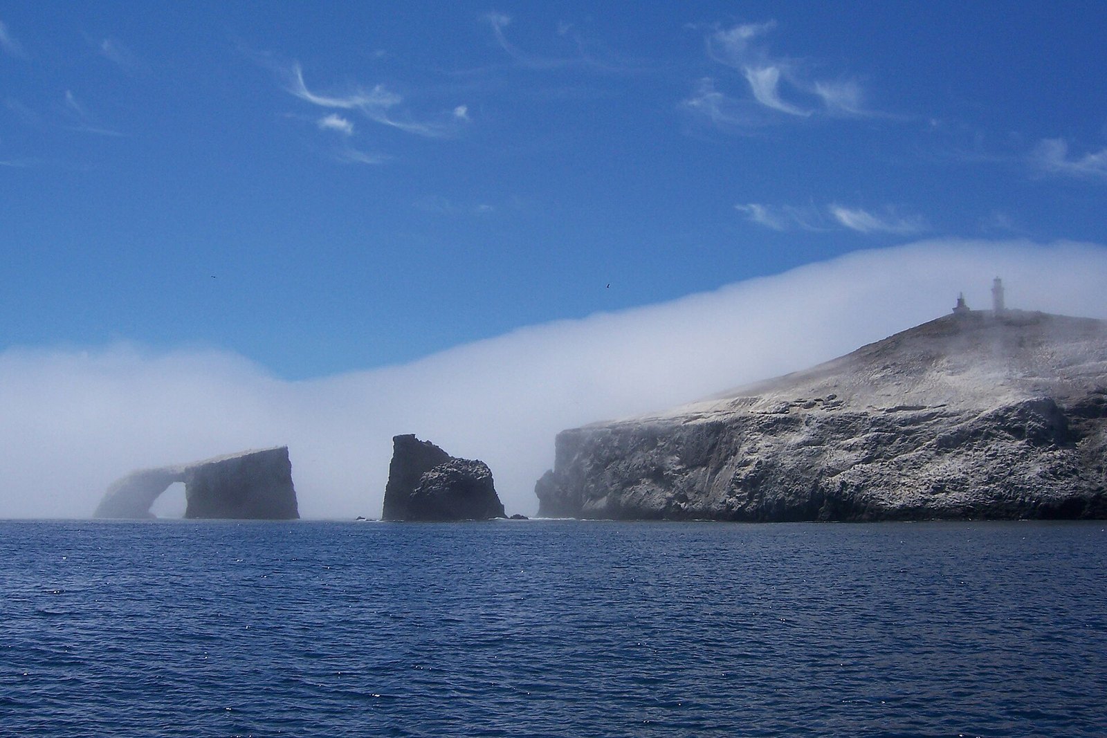 A rock arch and a sea stack off the end of a pale cliffed island topped by a lighthouse, with a bank of fog rolling over blue water under a blue sky