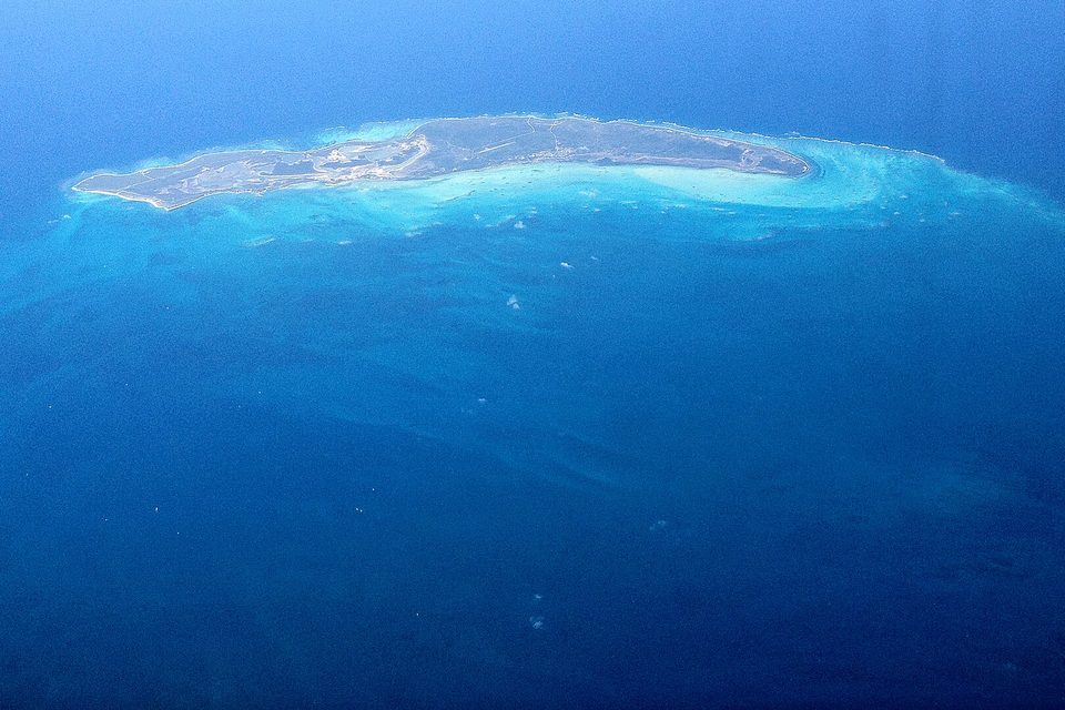 A long, flat, grey-green island seen from high above, with pale turquoise shallows and a reef curving around it in deep-blue ocean