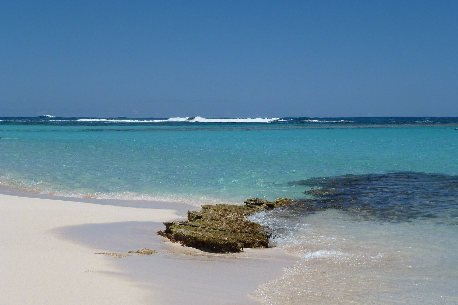 Powder-white sand curves past a low shelf of coral rock into clear turquoise shallows, with the white surf line of the outer reef across the horizon under a deep-blue sky