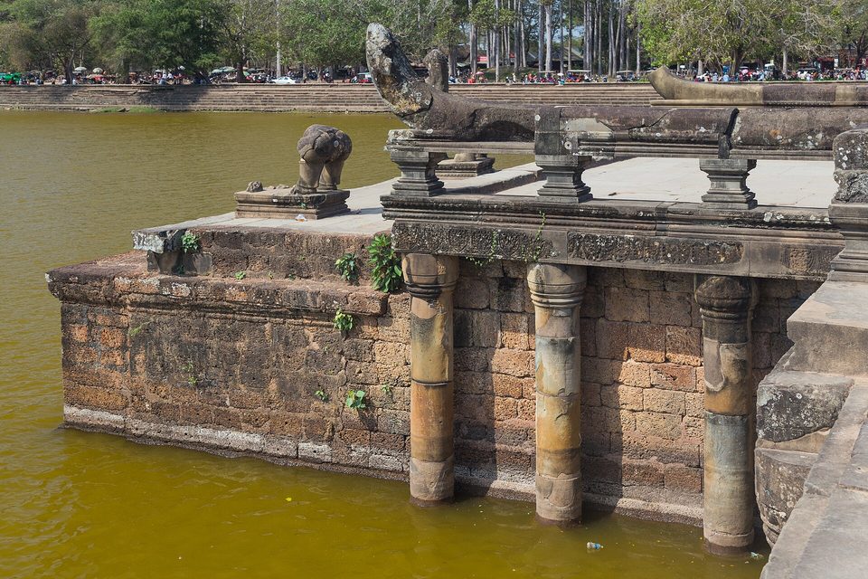 A brick and sandstone terrace with a carved balustrade rising from green moat water, trees and a far bank beyond