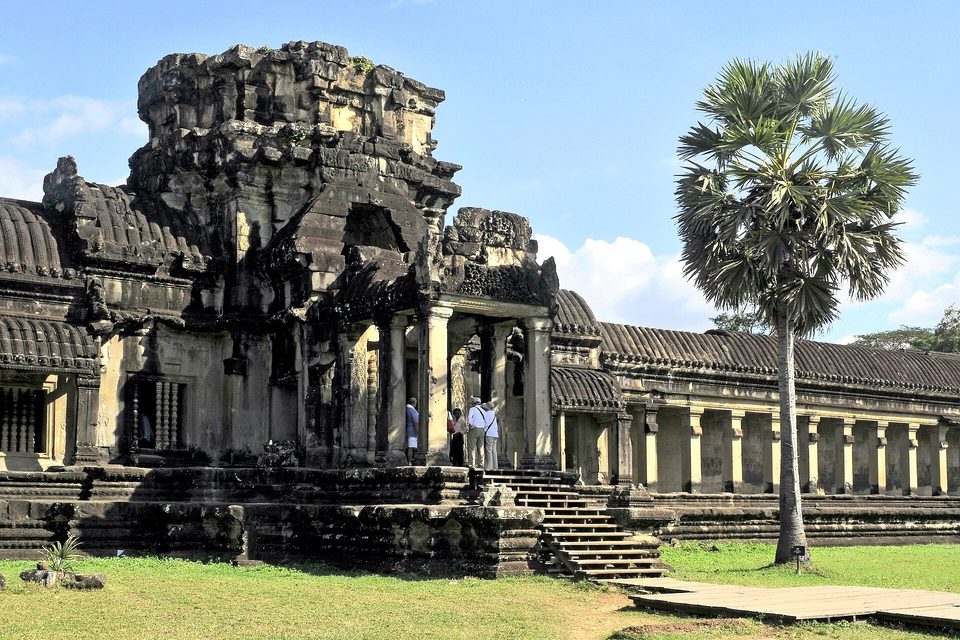 A dark stone gateway tower with a lawn in front and a pillared gallery running away to the right under a blue sky