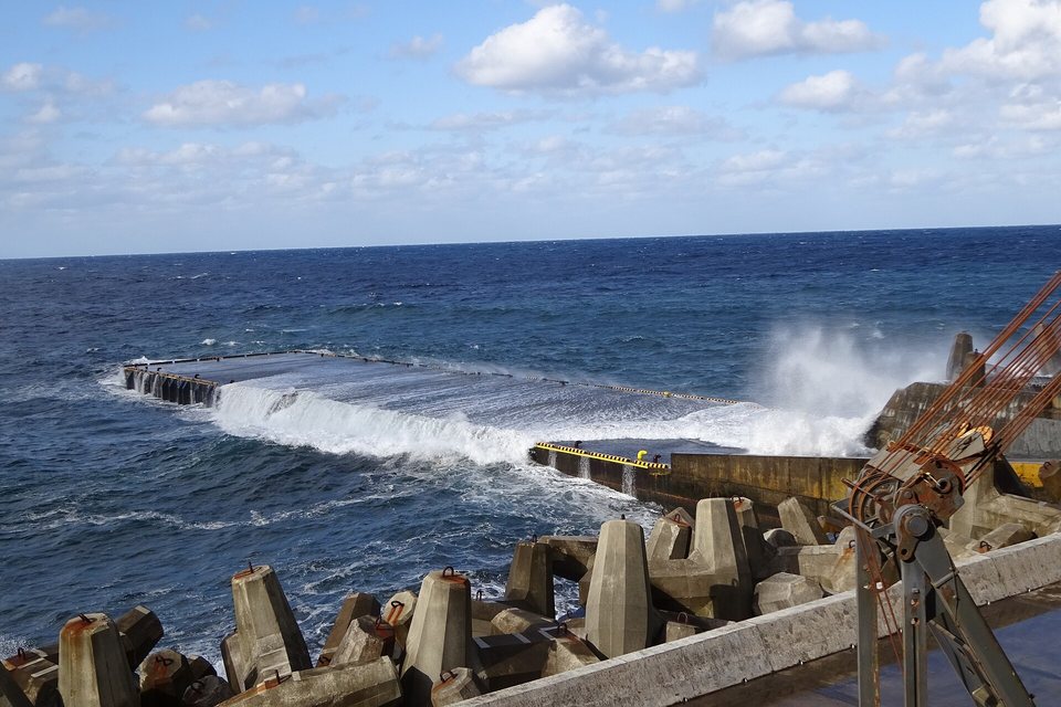 A wave washing white over a low concrete wharf in a rough blue sea, concrete breakwater blocks in the foreground