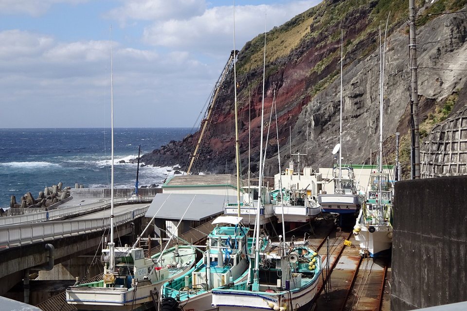 Small fishing boats hauled up on a steep slipway on rails, below a red and grey volcanic cliff, rough sea to the left
