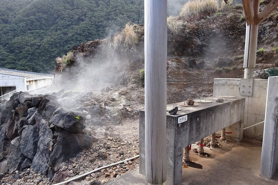 A concrete cooking bench with round lids over steam pipes, steam drifting from a bank of dark volcanic rocks beside it
