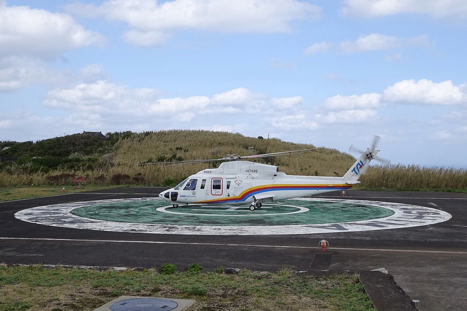 A white helicopter with a rainbow stripe parked on a green-and-white helipad, a grassy hill behind