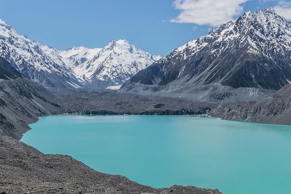 A turquoise lake beneath a wall of grey moraine, with snow-covered peaks beyond