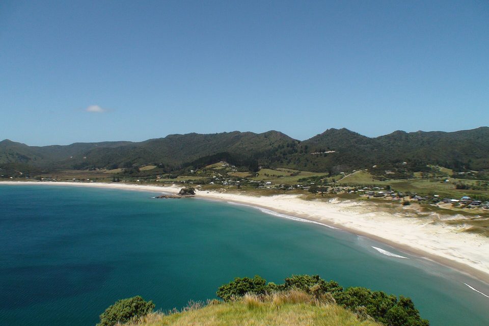 A long white surf beach curves beneath green, bush-covered hills, with a scatter of houses on the flat behind the dunes